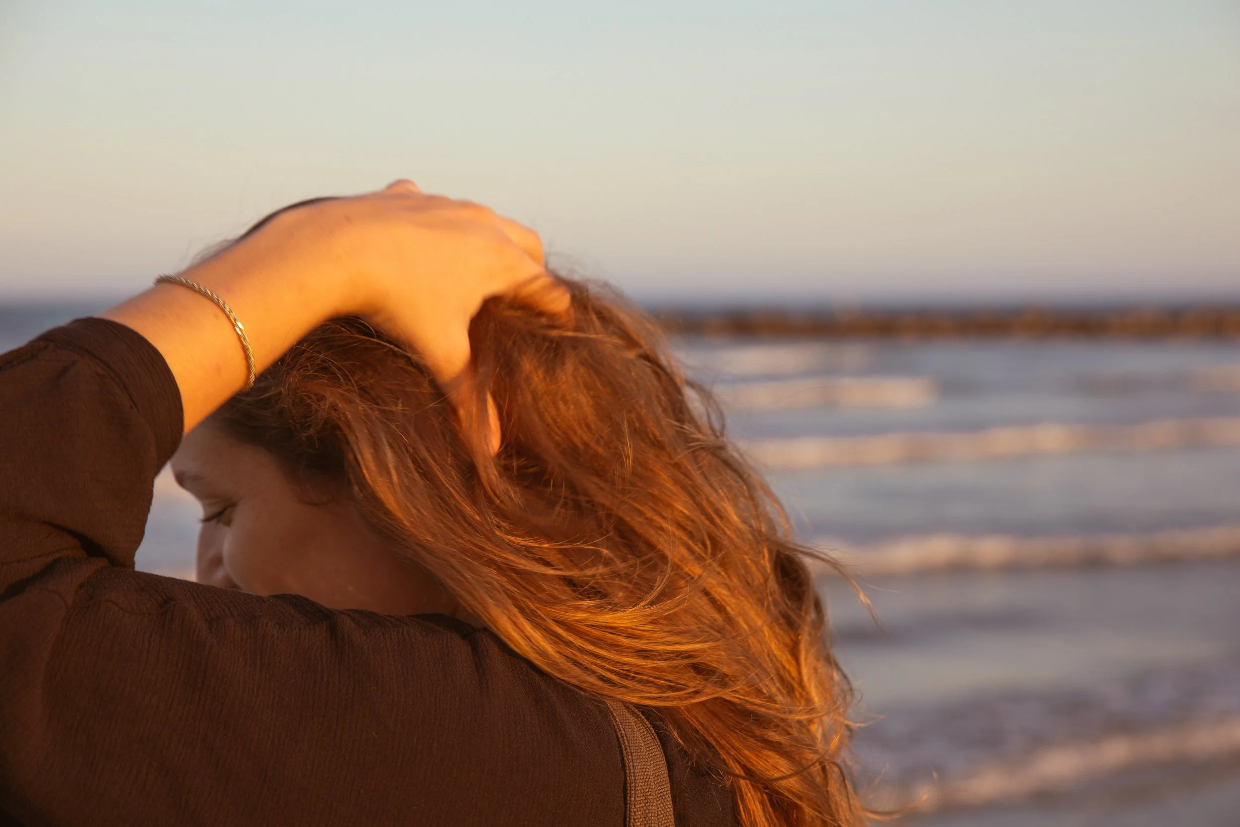 Une femme avec des cheveux roux, portant un bracelet en or, regarde vers la mer lors du coucher de soleil.