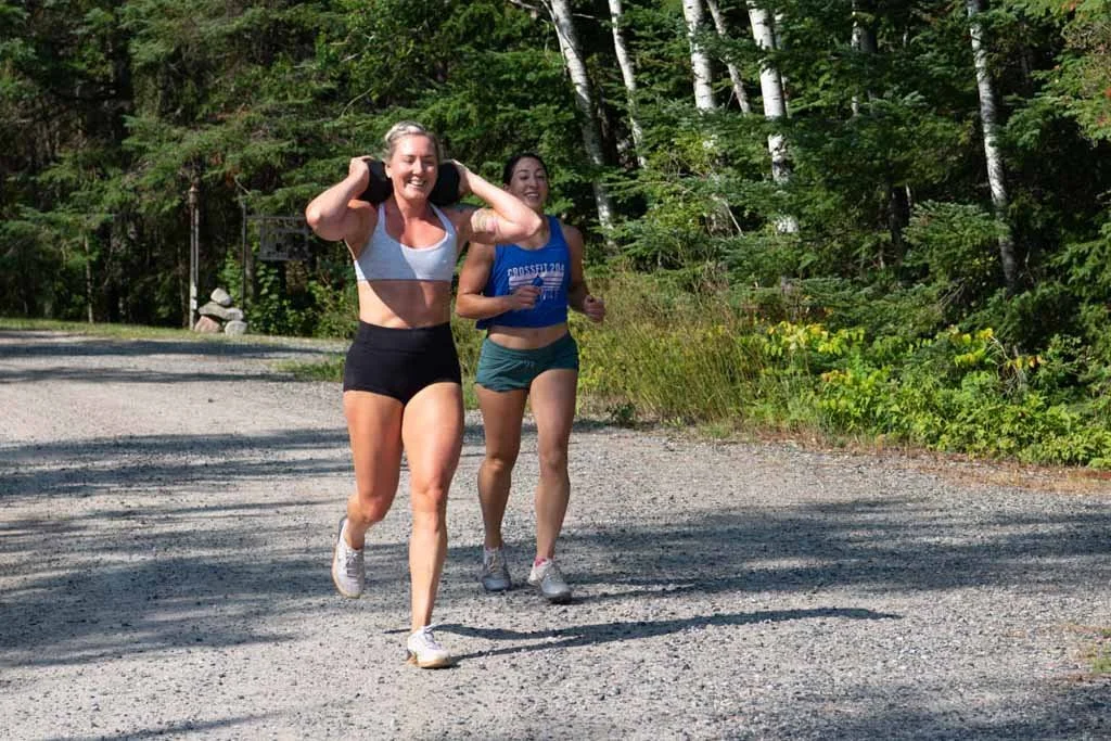 Two women running on a gravel trail in a wooded area, smiling and enjoying exercise together.