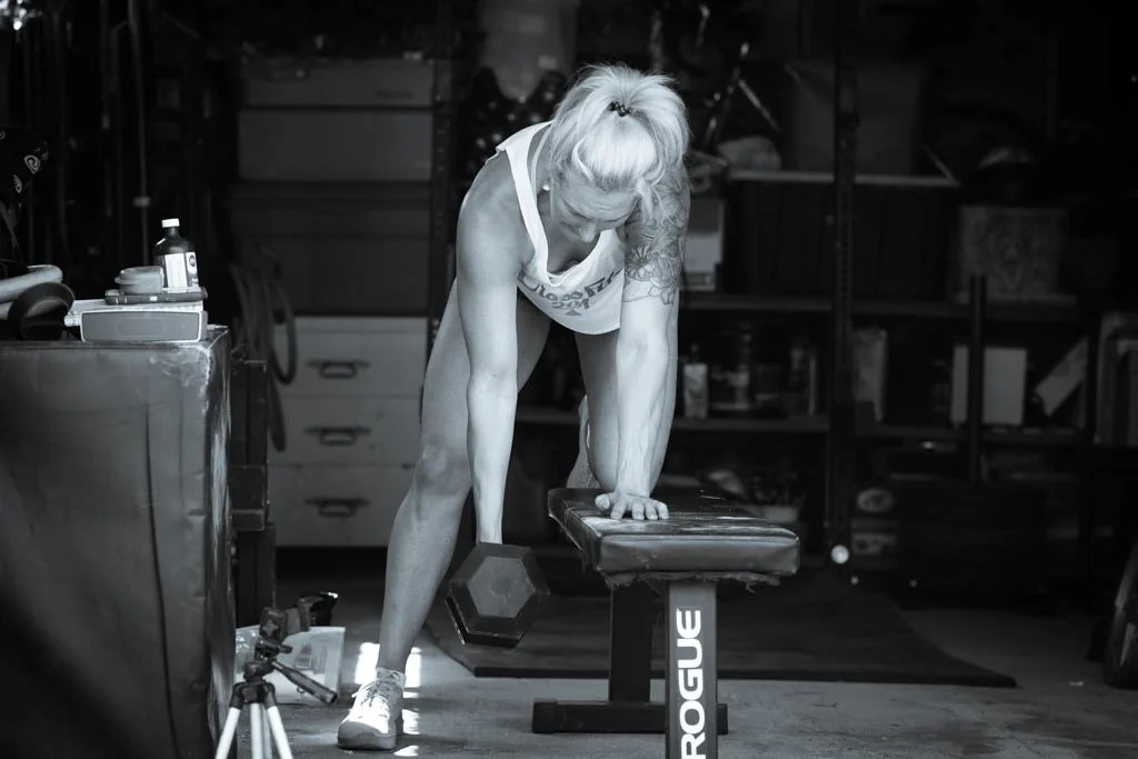 A woman with blonde hair in a ponytail, wearing a tank top and shorts, is exercising in a gym. She is using a bench and holding a dumbbell with one hand, leaning on the bench, with her other hand on her knee, in a workout routine.