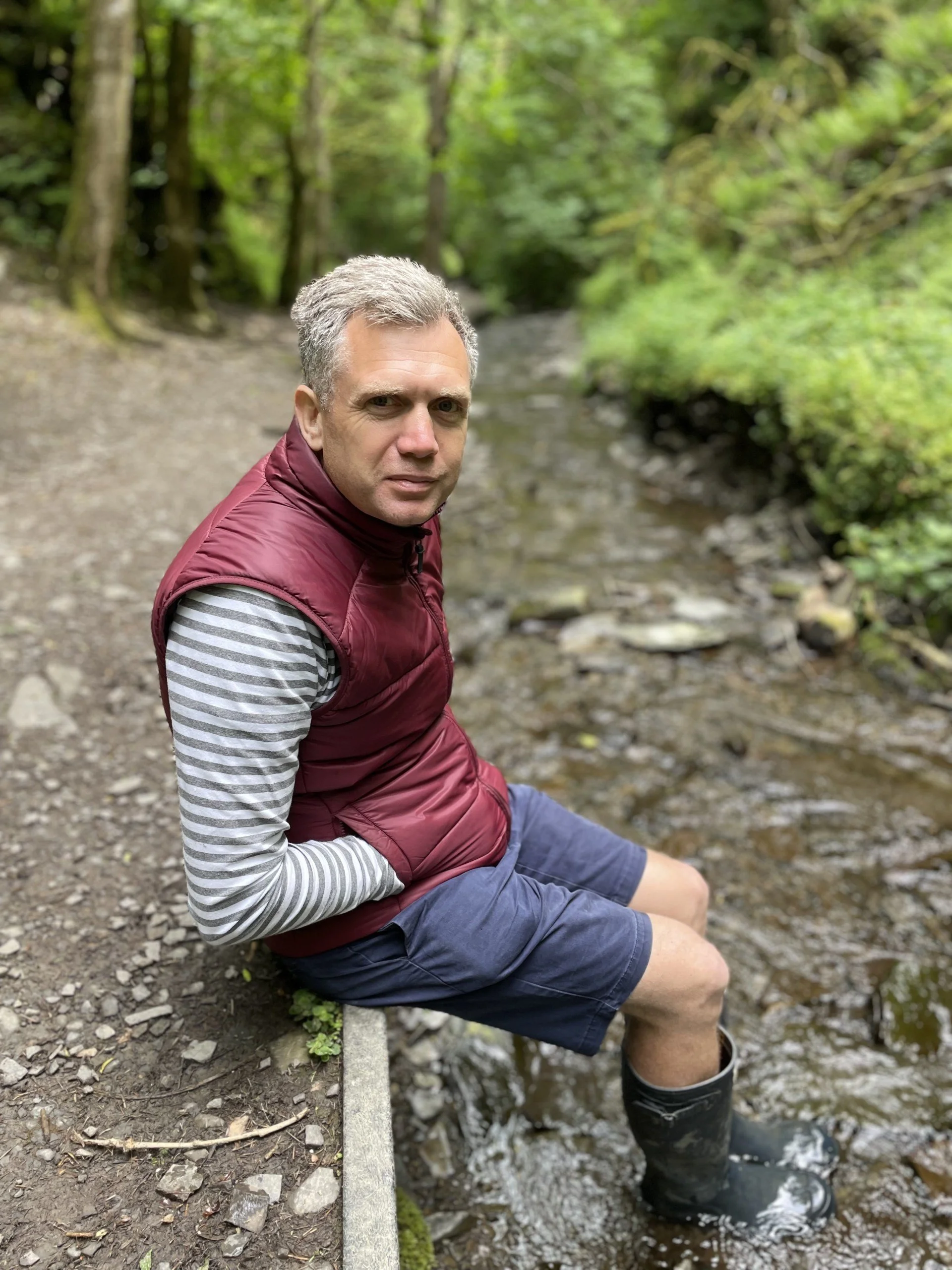 A man with gray hair sitting by a stream in a wooded area, wearing a maroon vest over a striped long-sleeve shirt, shorts, and rubber boots.