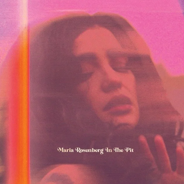 Photograph of a woman with long hair looking into a mirror, with a pink background. The text overlay reads "Maria Rosenberg In The Pit."