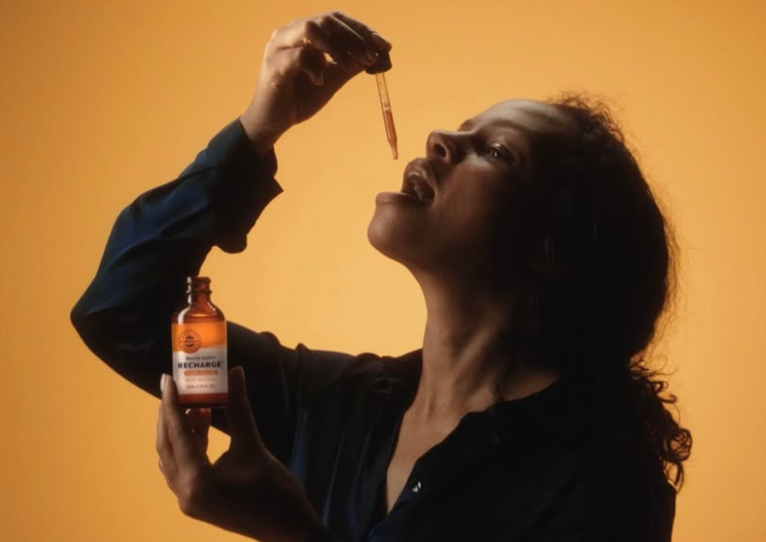 A woman with curly hair in a black shirt holding a small bottle labeled 'Recharge' and using a dropper to put liquid into her mouth, with a yellow background.