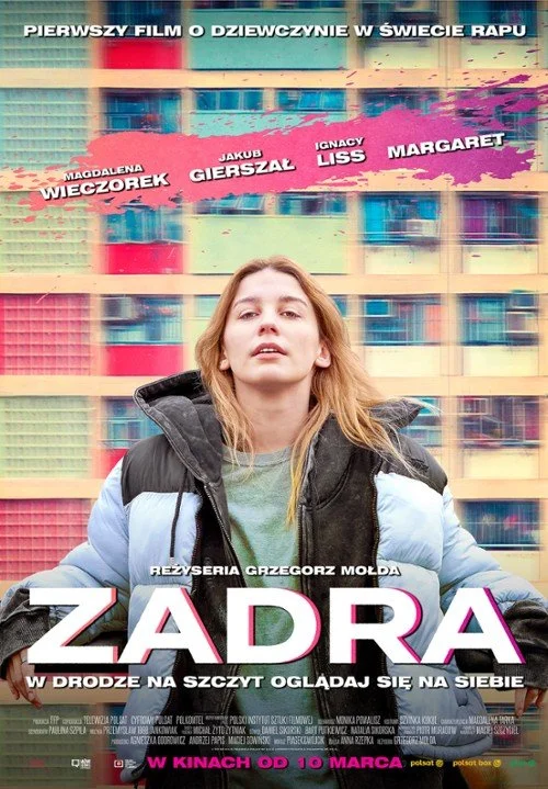 A woman standing in front of a colorful, geometric-patterned building, wearing a puffy jacket and looking up with a serious expression.