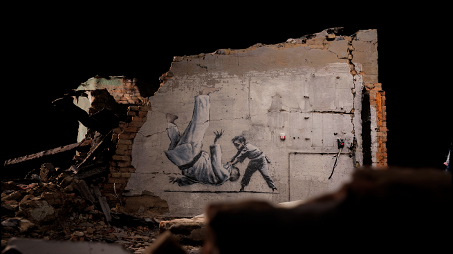 Graffiti of two people, one falling and the other reaching out, painted on a damaged wall surrounded by debris and rubble.