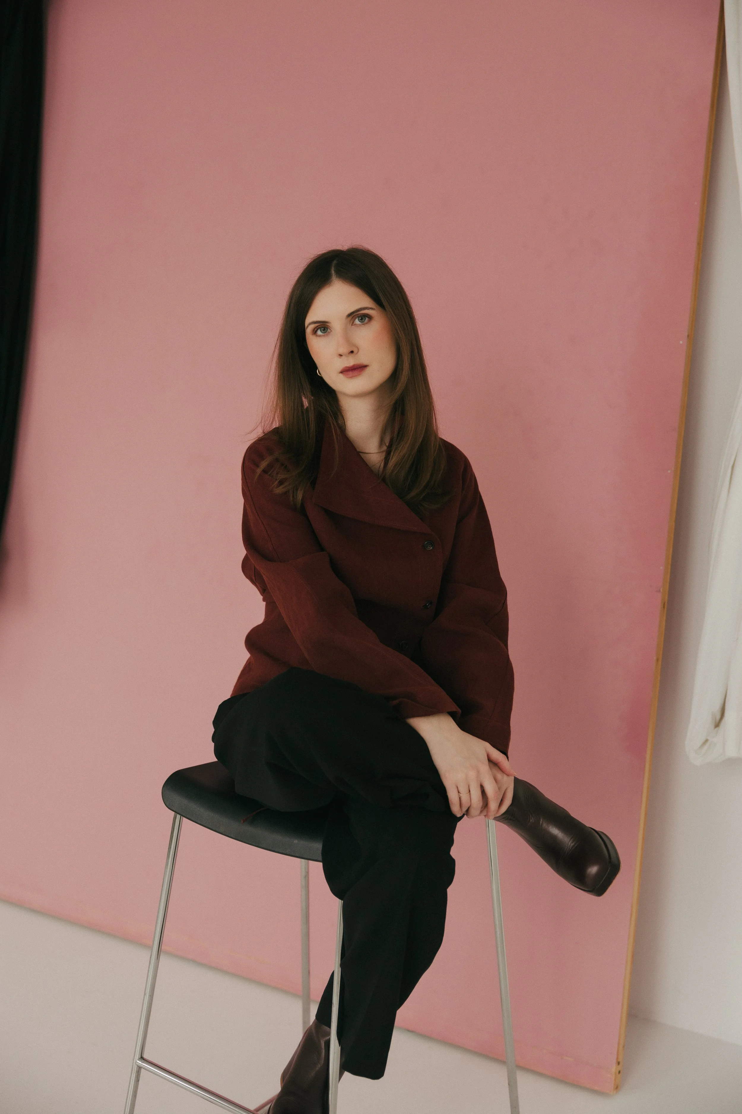 A young woman with long brown hair and fair skin, wearing a dark red blazer and black pants, is sitting on a black chair in front of a pink backdrop. She is looking directly at the camera with her right arm resting on her lap and her left arm relaxed, with one leg crossed over the other, wearing dark brown boots.