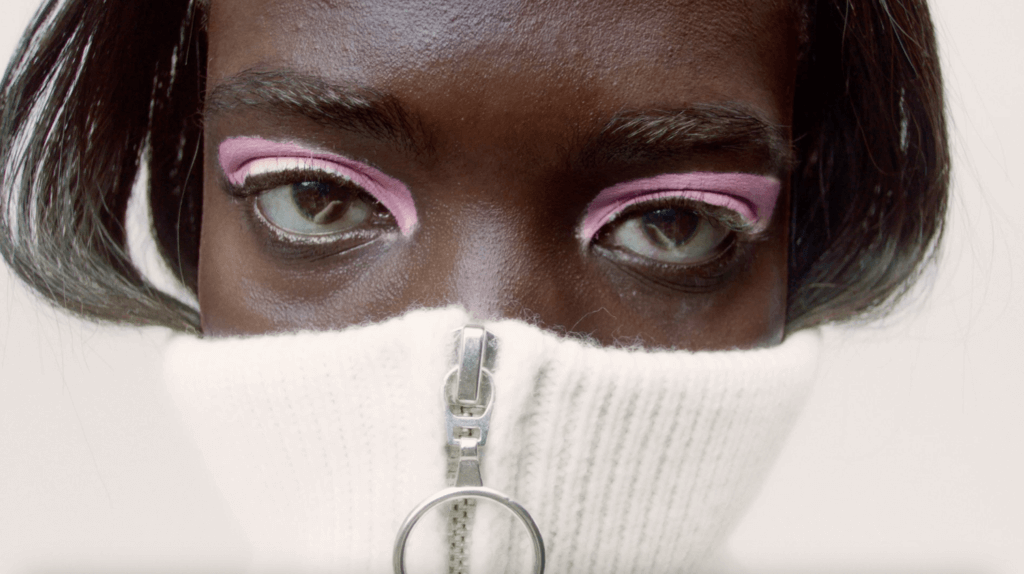 Close-up of person with dark skin wearing eye makeup with pink eyeshadow and a white zippered turtleneck covering lower face.