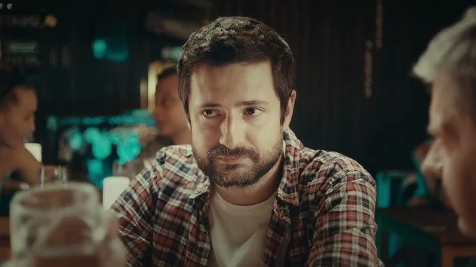 A man with dark hair and beard wearing a plaid shirt sitting at a bar or restaurant.