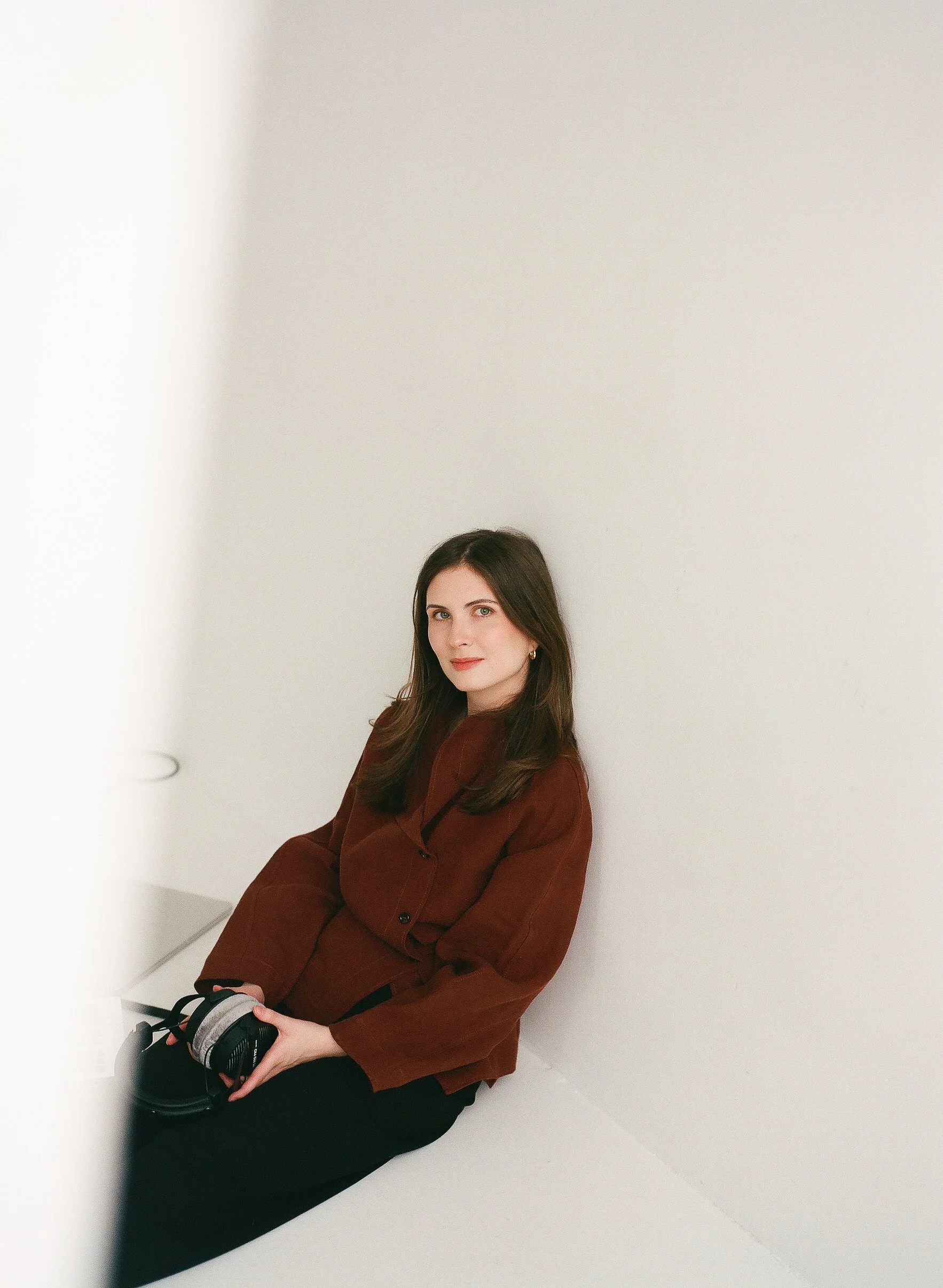 A woman with long brown hair sitting against a white wall, wearing a brown blazer and black pants, holding a pair of headphones in her hands, looking directly at the camera.