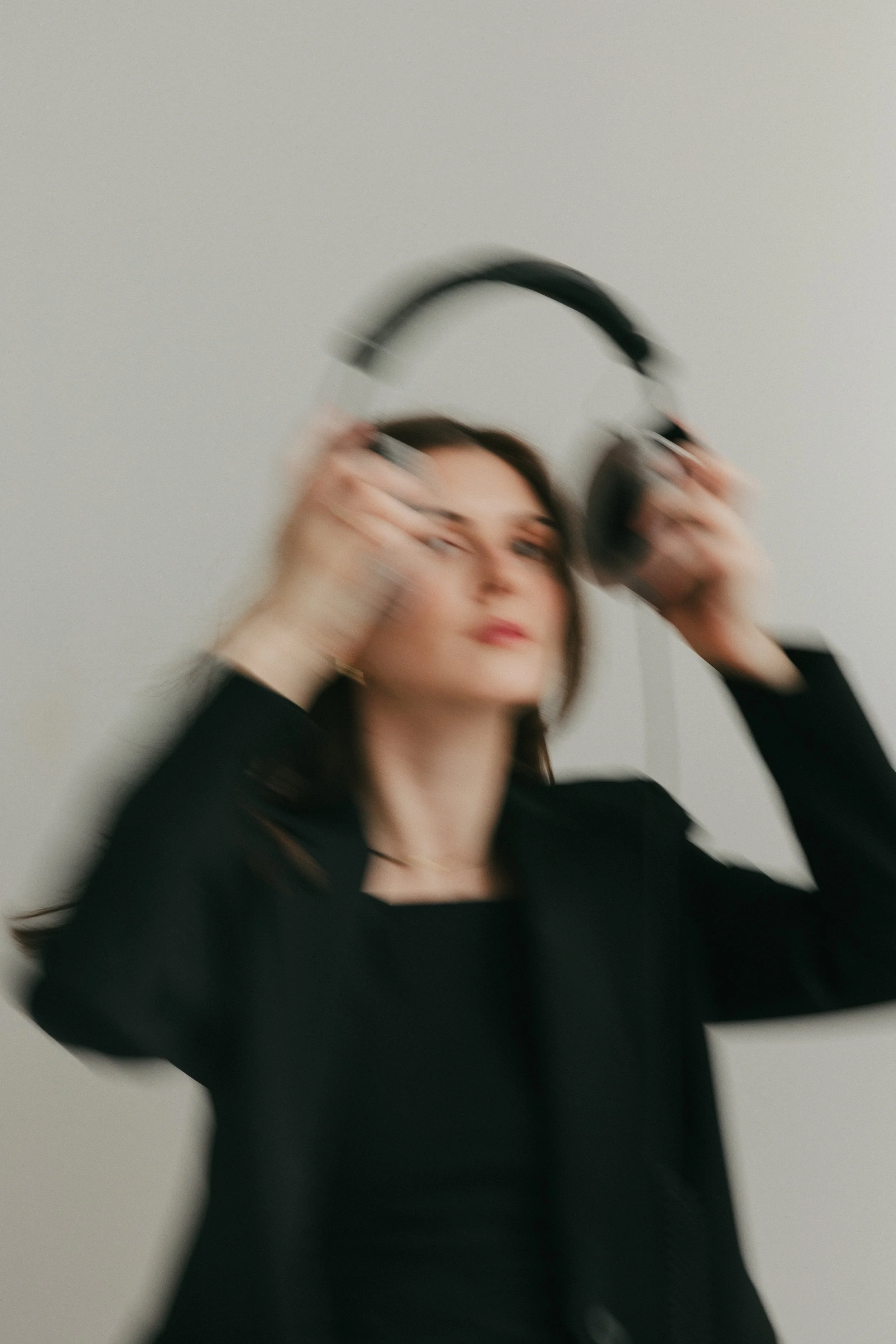 A woman with dark hair, wearing a black top, is holding black headphones to her ears against a plain light-colored background.