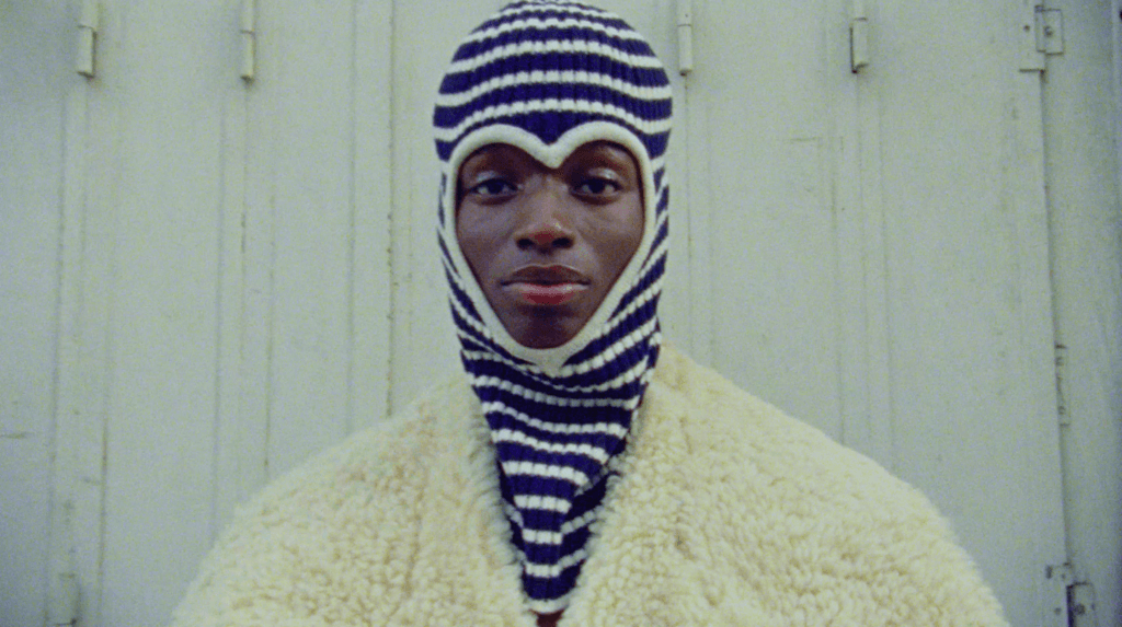 Person wearing a blue and white striped knit hat that covers their head and neck, and a cream-colored fluffy coat, standing in front of a light-colored wooden wall.