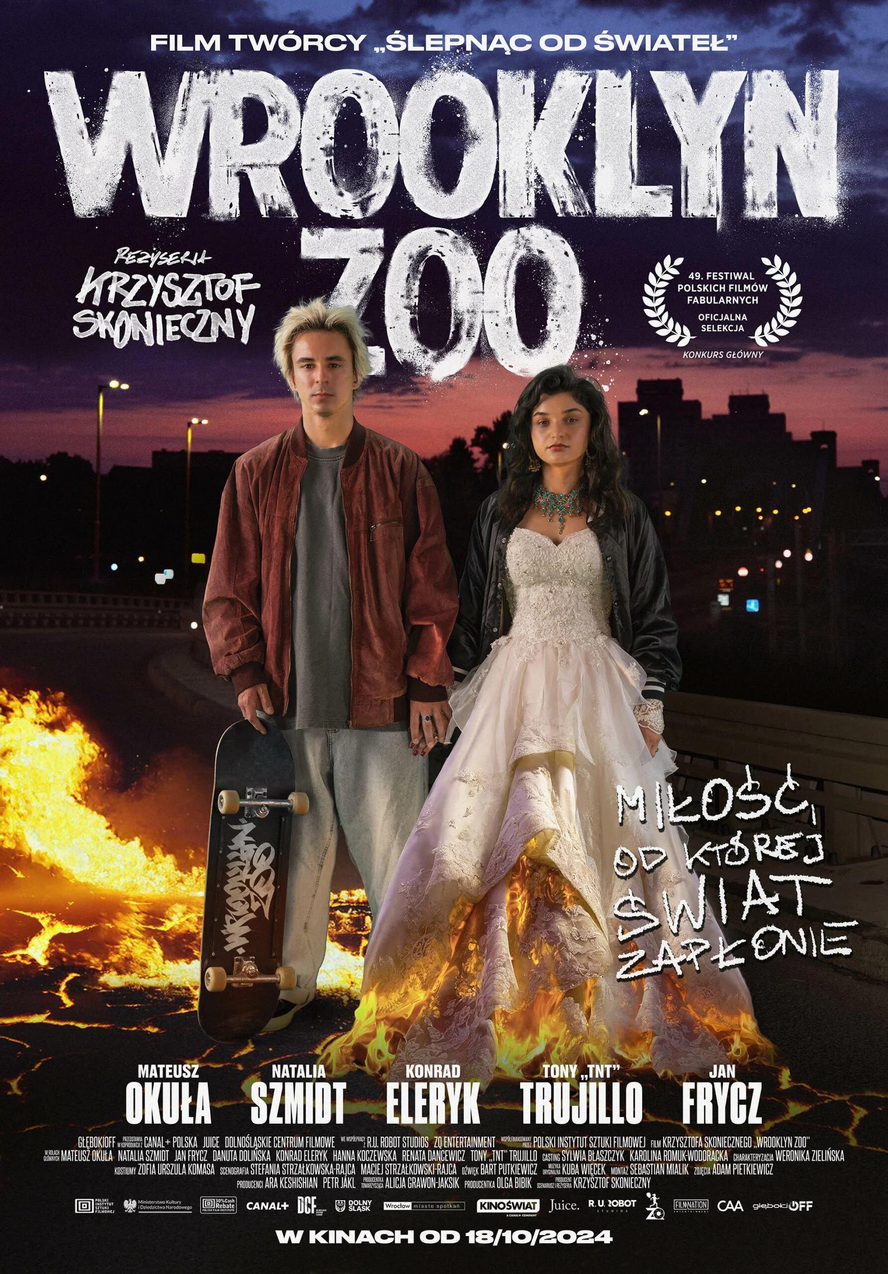 Poster for the film "Wroclaw 700" featuring a young man with blond hair holding a skateboard and a young woman in a white dress with a black jacket, standing against a background of a city skyline, a sunset sky, flames, and explosion. The poster includes text in Polish about the film's creators, cast, and release date October 18, 2024.
