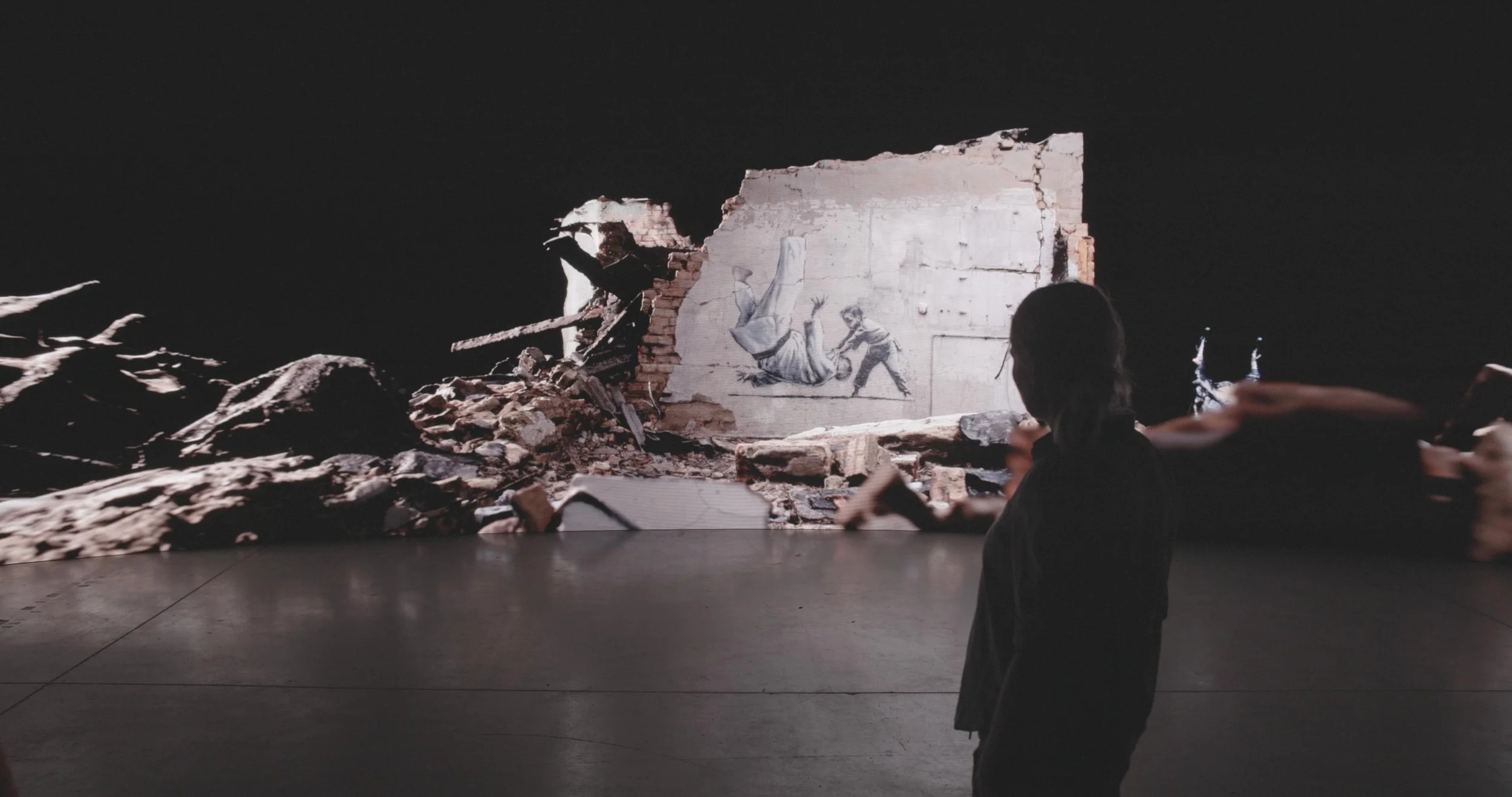 An art installation resembling a ruined wall with a mural of a person falling, surrounded by rubble, with onlookers observing.