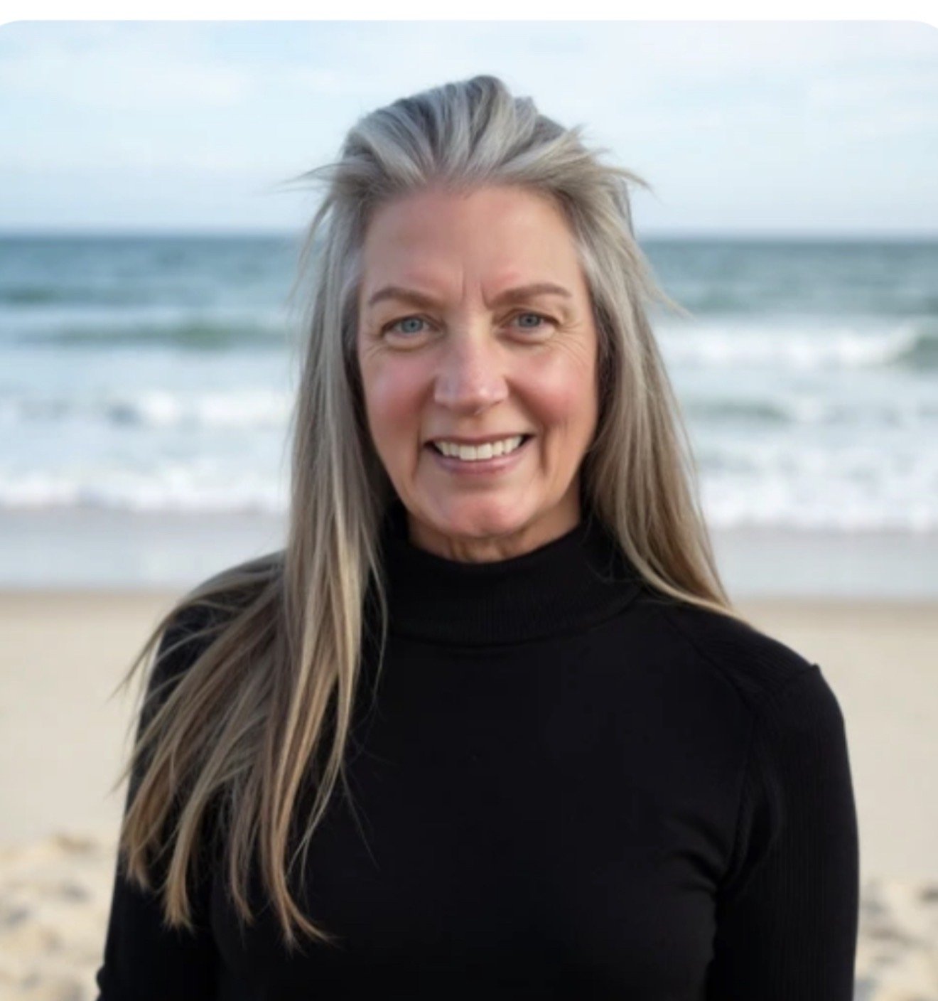 Smiling woman with long blonde hair in a black turtleneck, standing on a beach with the ocean and waves in the background.