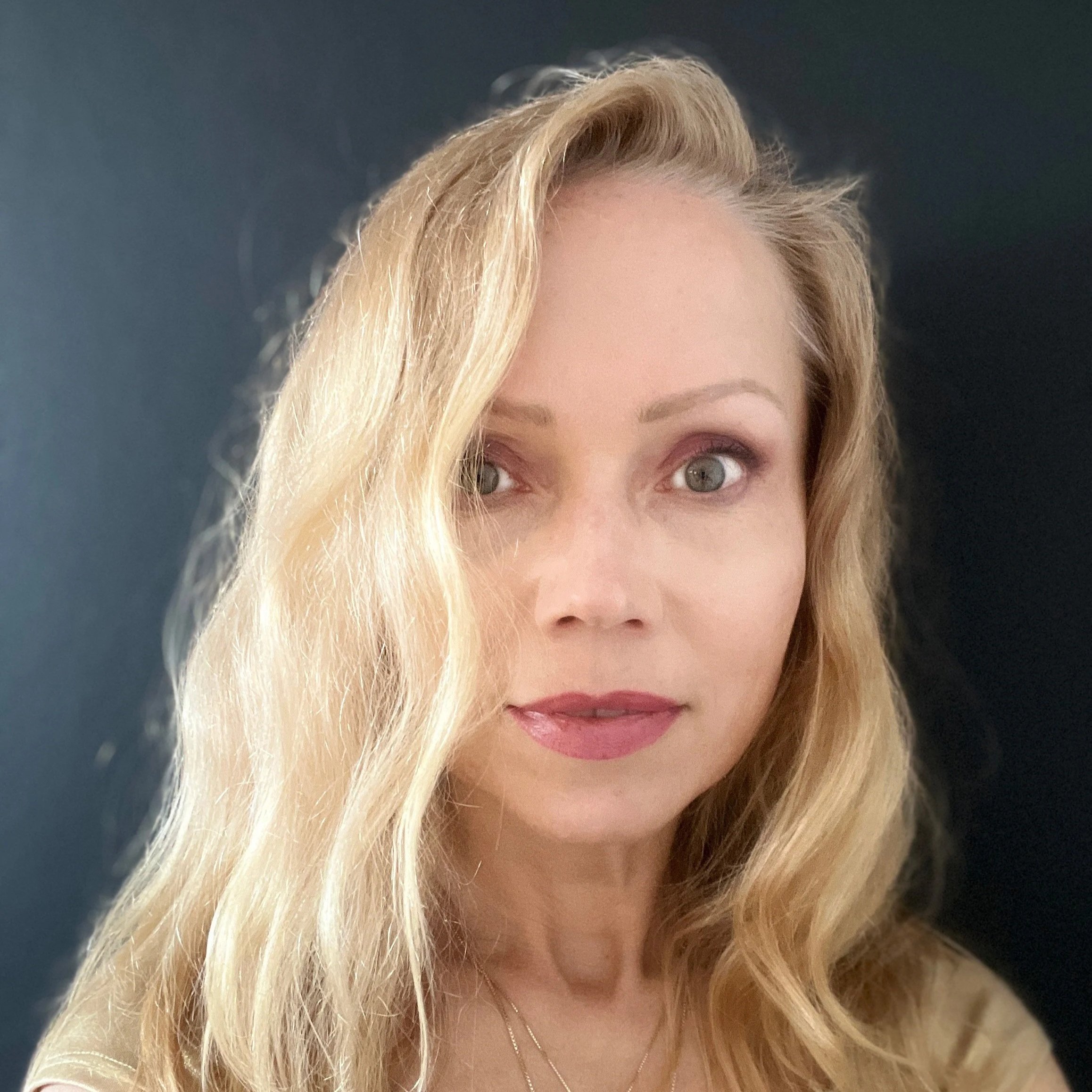 Close-up of a woman with long, wavy blonde hair, light skin, and blue eyes, looking at the camera with a slightly open mouth, wearing makeup and a gold necklace, against a dark background.