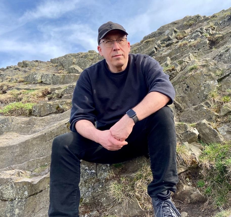 A man wearing a black cap, glasses, black sweatshirt, and black pants sitting on rocky terrain outdoors, with a cloudy sky in the background.