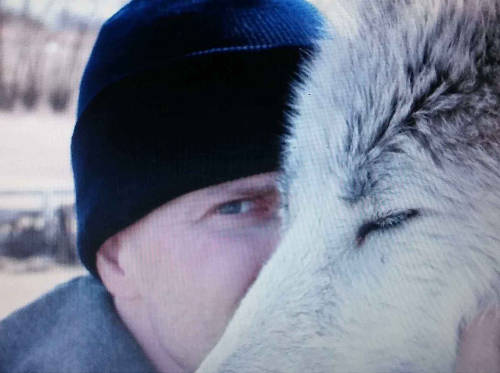 A person wearing a black beanie hat, with one eye visible, embracing a large, fluffy animal, possibly a dog or a wolf.