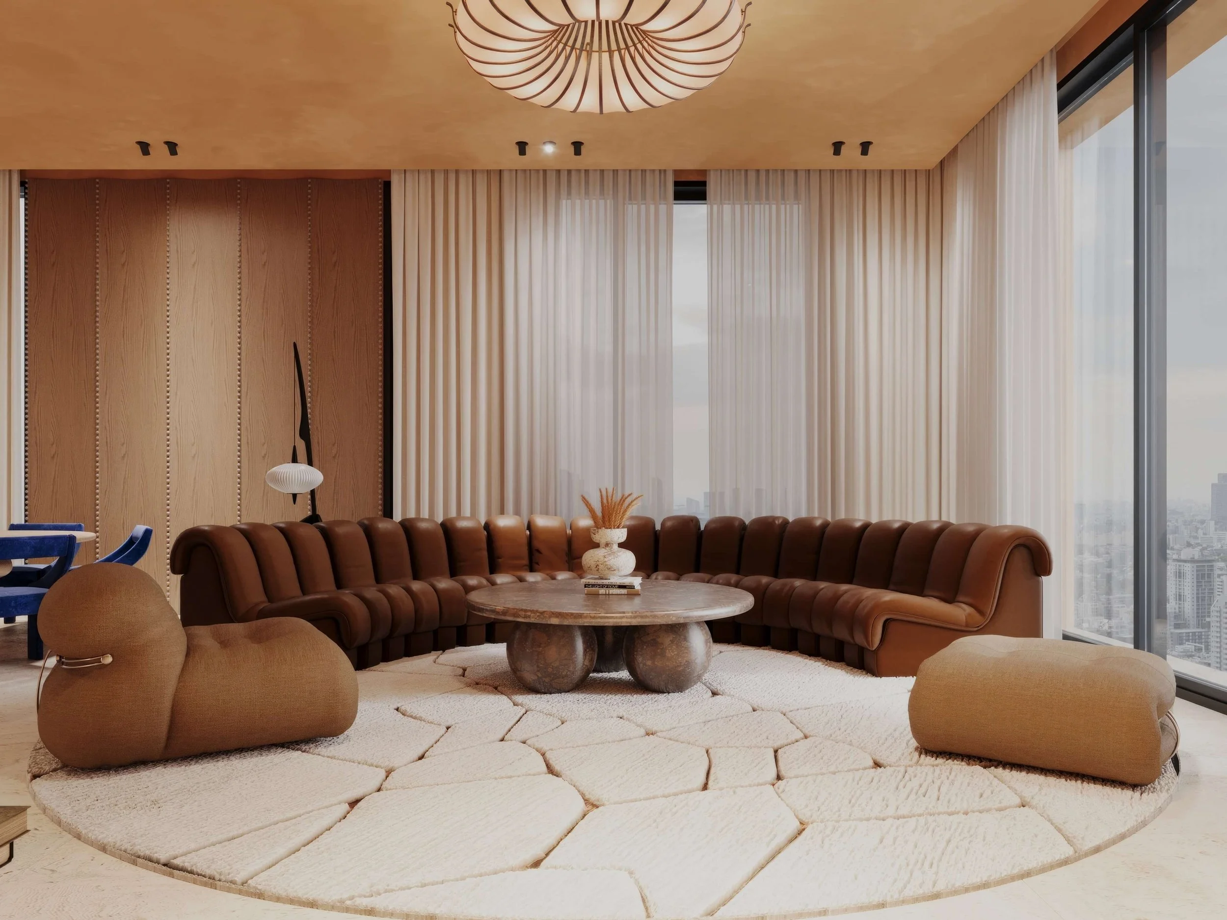Modern living room with a large L-shaped brown leather sofa, a round coffee table with a vase and decorative items, beige and brown furniture, large windows with sheer curtains, and city skyline view.