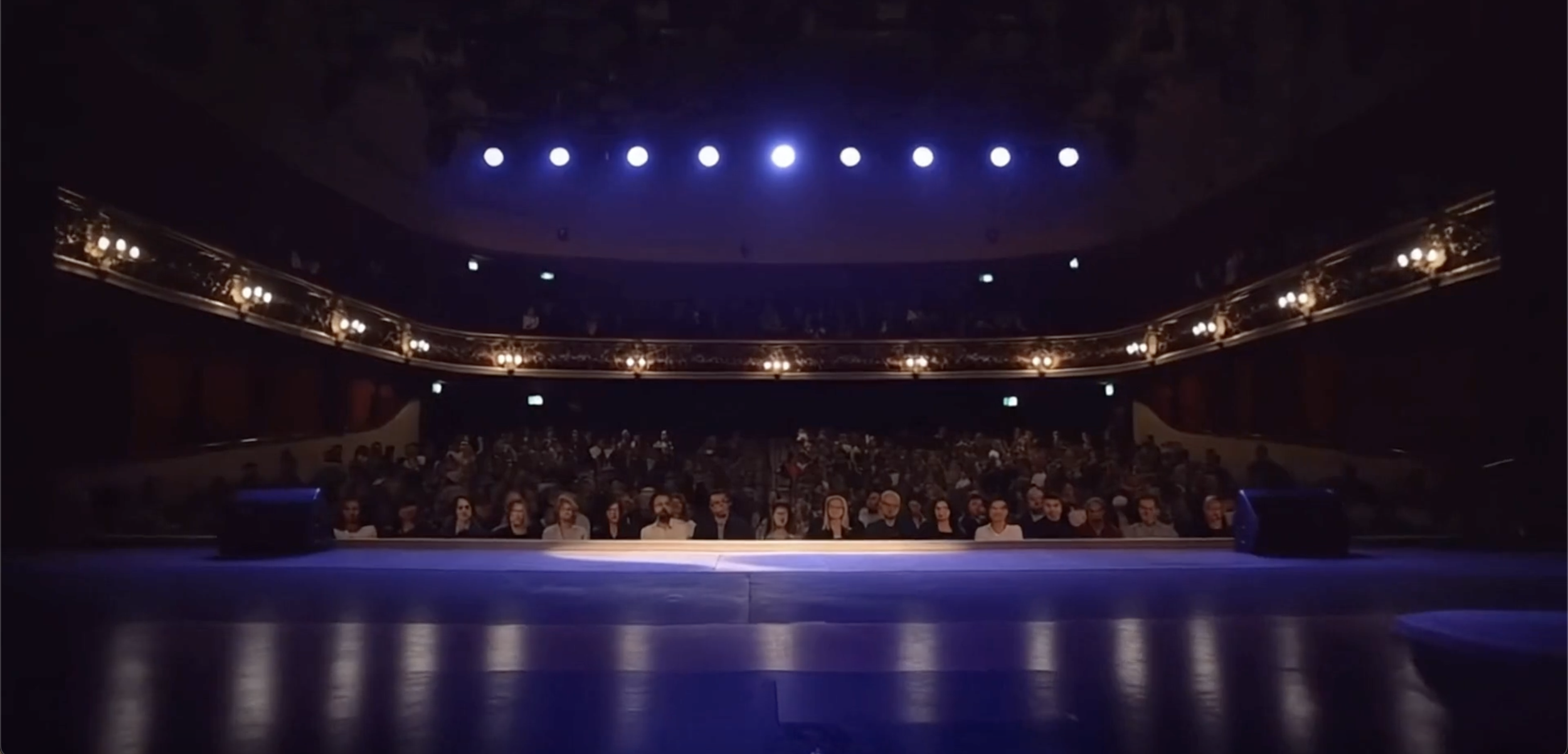 Une scène avec un public, la caméra placée sur l'estrade avance lentement vers les spectateurs 