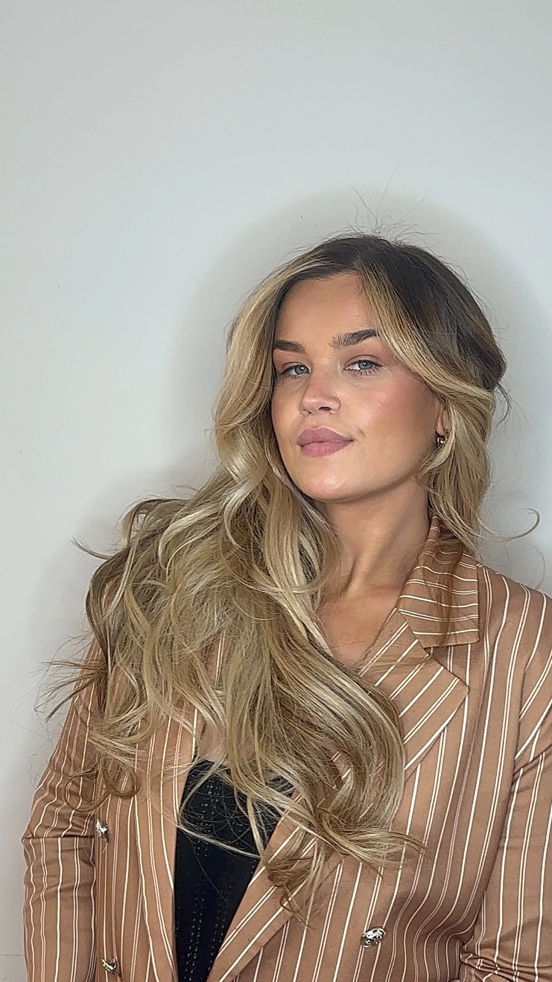 Porträt einer jungen Frau mit langen, blonden, lockigen Haaren, blaue Augen und braunem Lippenstift, trägt ein braun gestreiftes Hemd gegen eine weiße Wand.