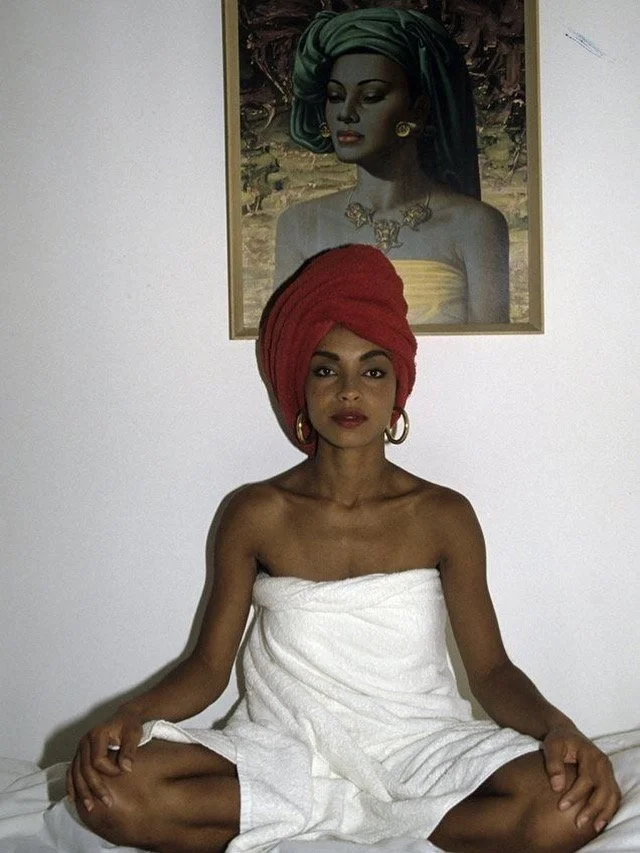 Frau sitzend in weißen Handtüchern, mit rotem Turban auf dem Kopf, in einem Raum mit einem Wandbild einer weiteren Frau im Hintergrund.