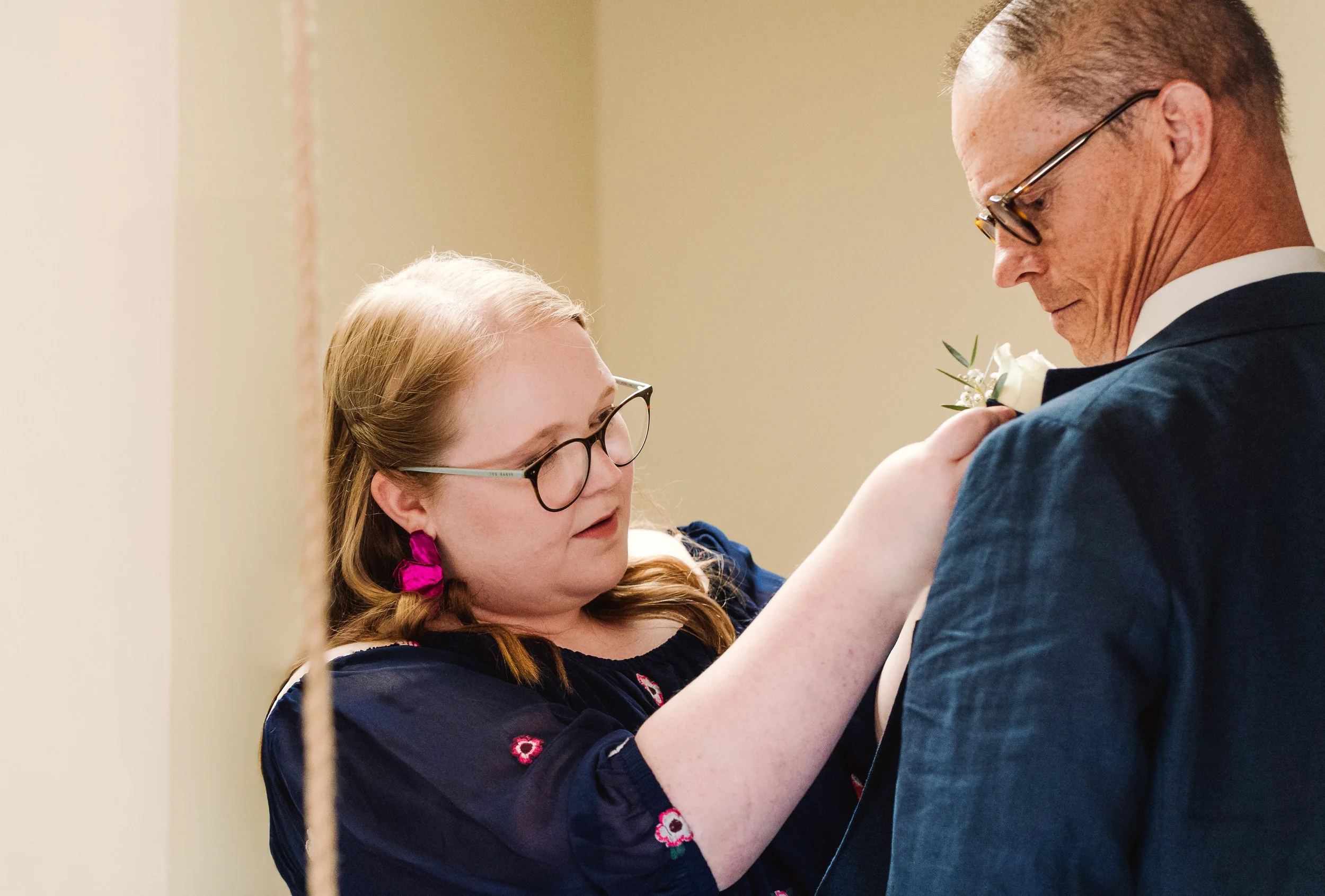 A girl with glasses and pink earrings pins a white boutonniere onto a man's blue suit jacket at a wedding.