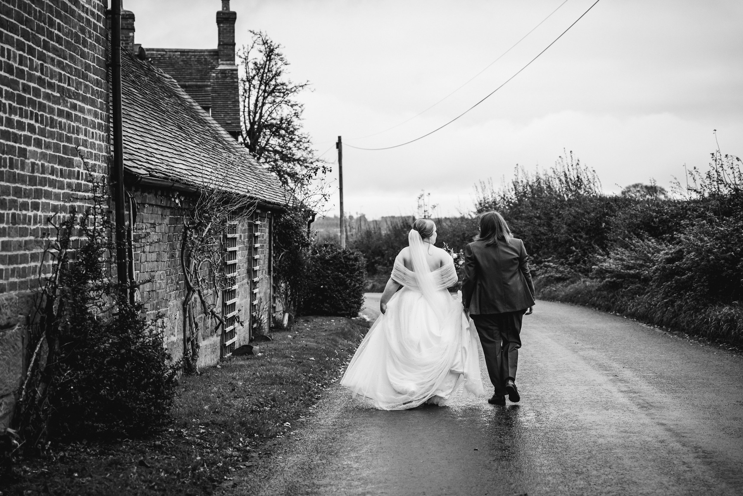 A couple dressed in wedding attire walking down a rural road, with a bride in a white gown and veil, and a bride in a suit, holding hands.