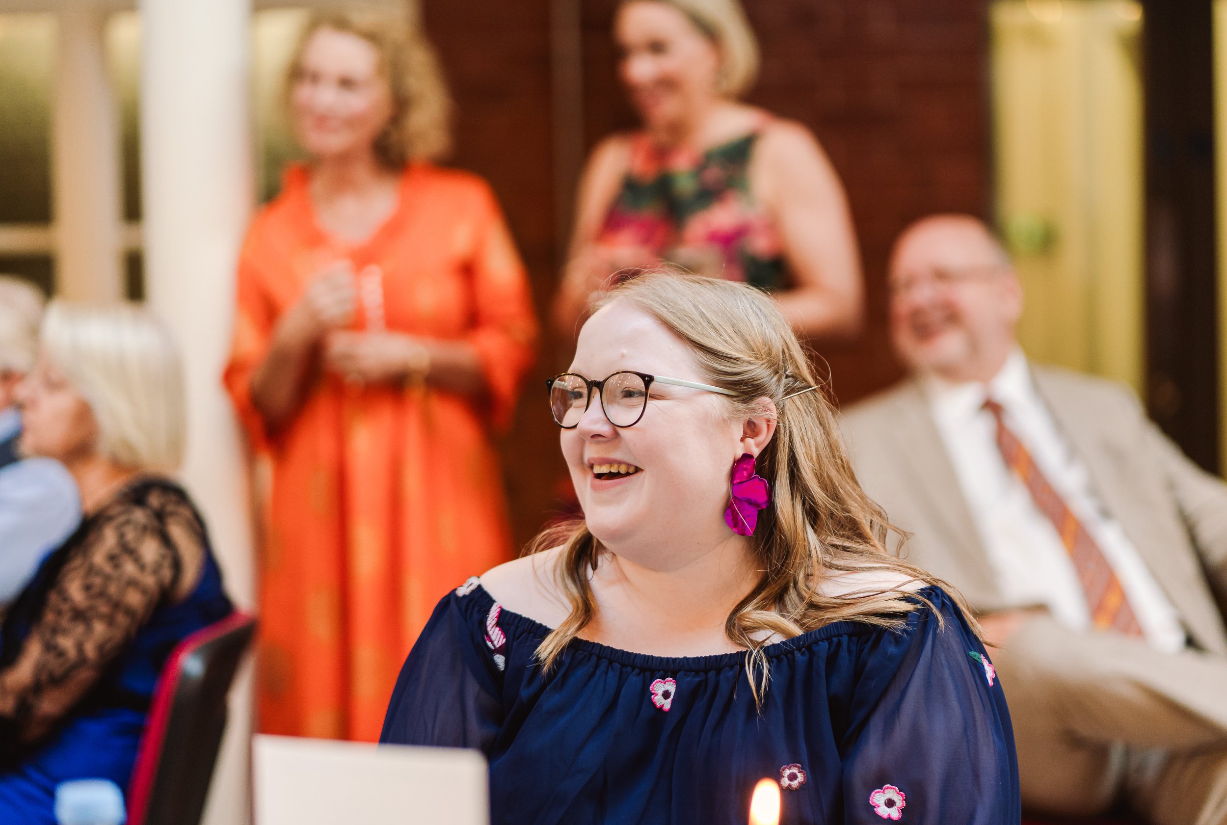 A woman with glasses and pink earrings smiling at a wedding, with several people in the background.