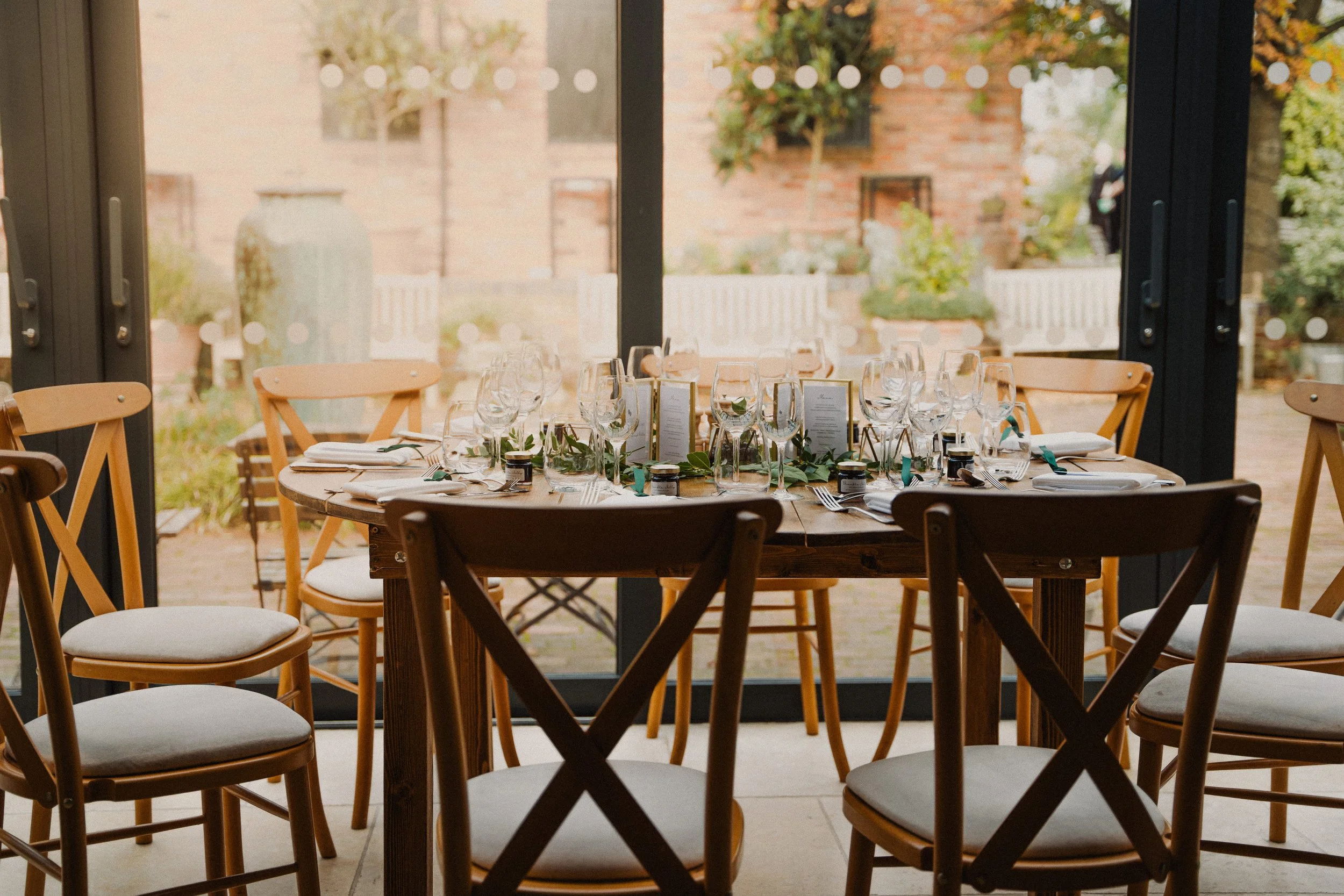 Round wooden table set for a formal event with multiple wine glasses, cutlery, and place settings, surrounded by wooden chairs, near a large glass door with a garden view.