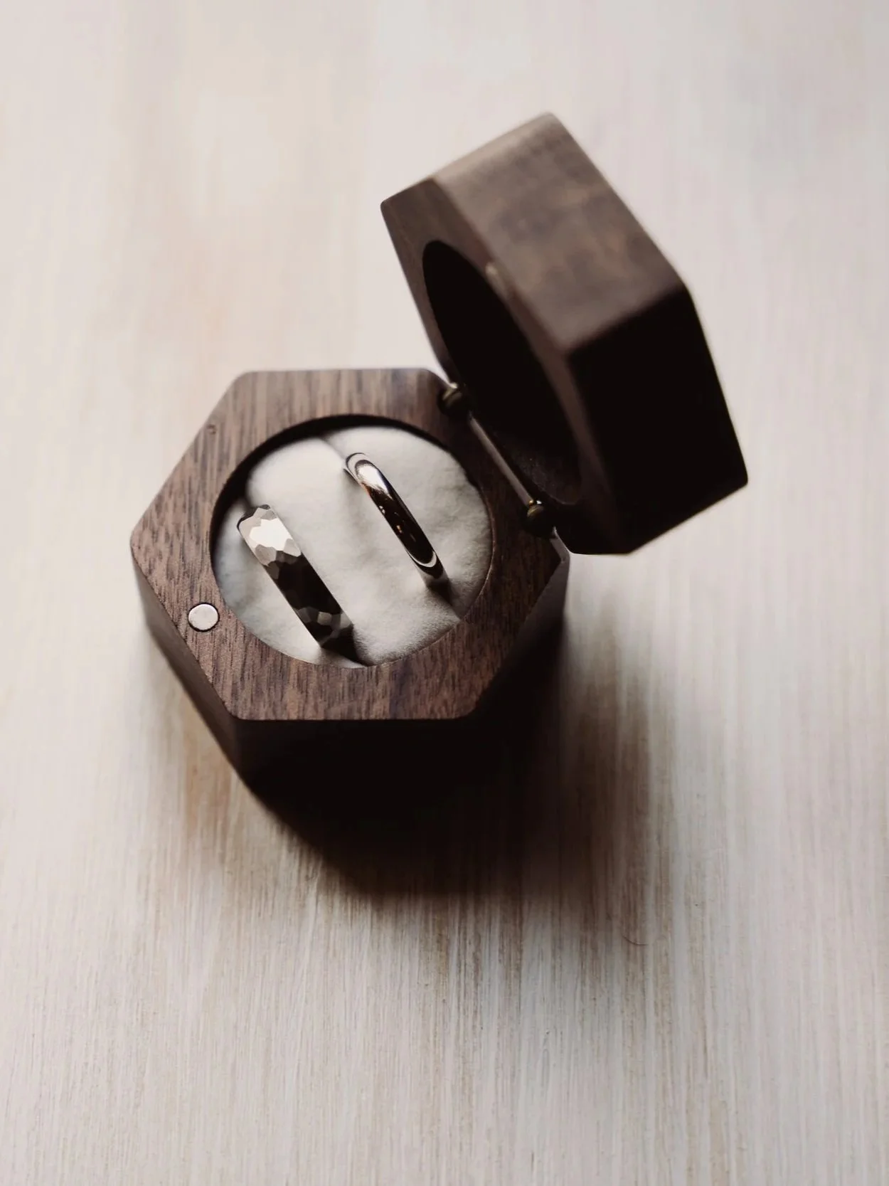 A wedding ring and a band inside a wooden ring box on a light wooden surface.