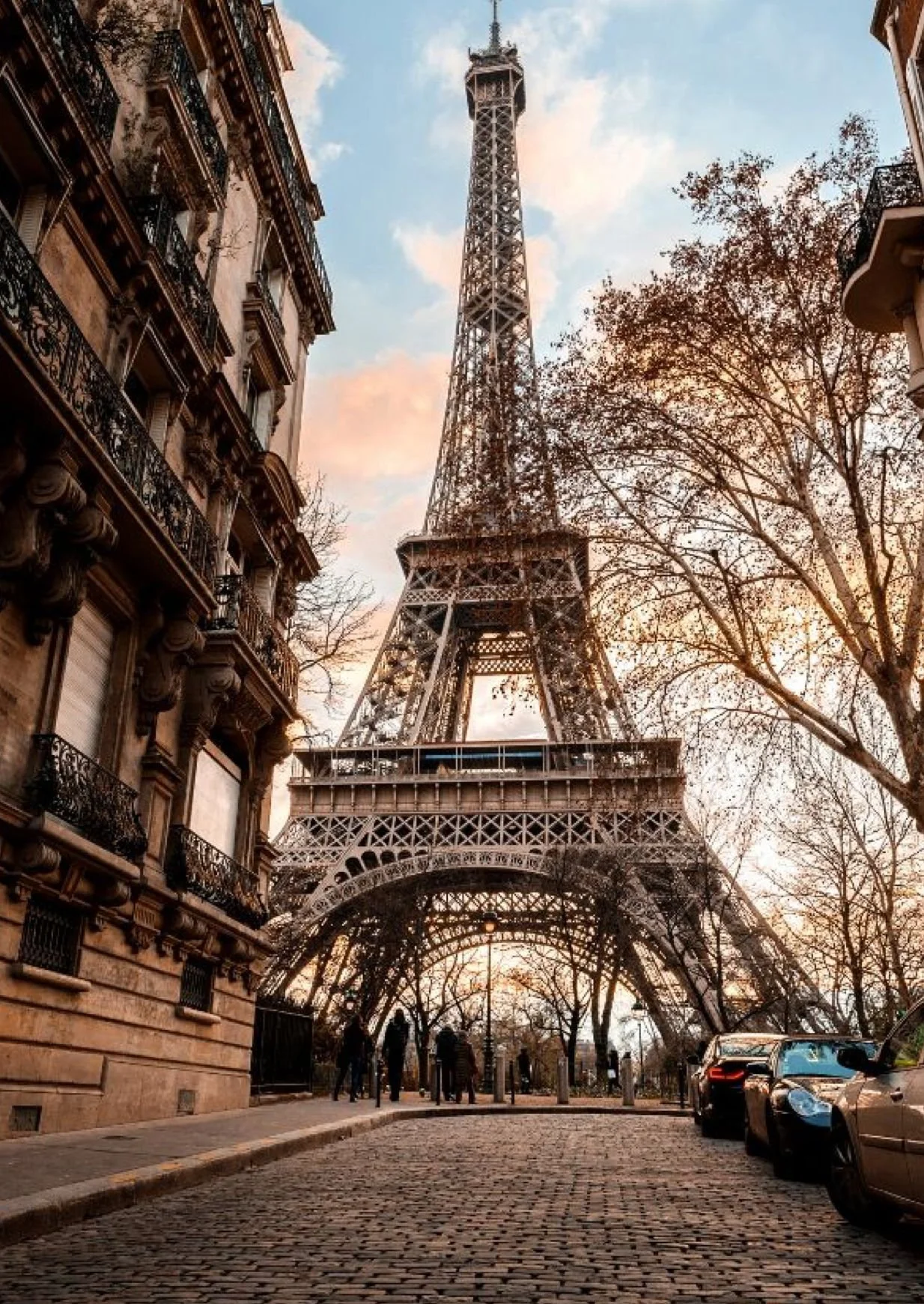 View of the Eiffel Tower in Paris during sunset, with trees and Parisian buildings on either side, and parked cars and people on the cobblestone street.