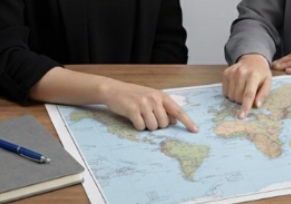 Two people pointing at a world map on a desk with a notebook and pen nearby.
