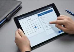 Person using a tablet to view a calendar and scheduling app on a desk with a closed laptop and a pen.