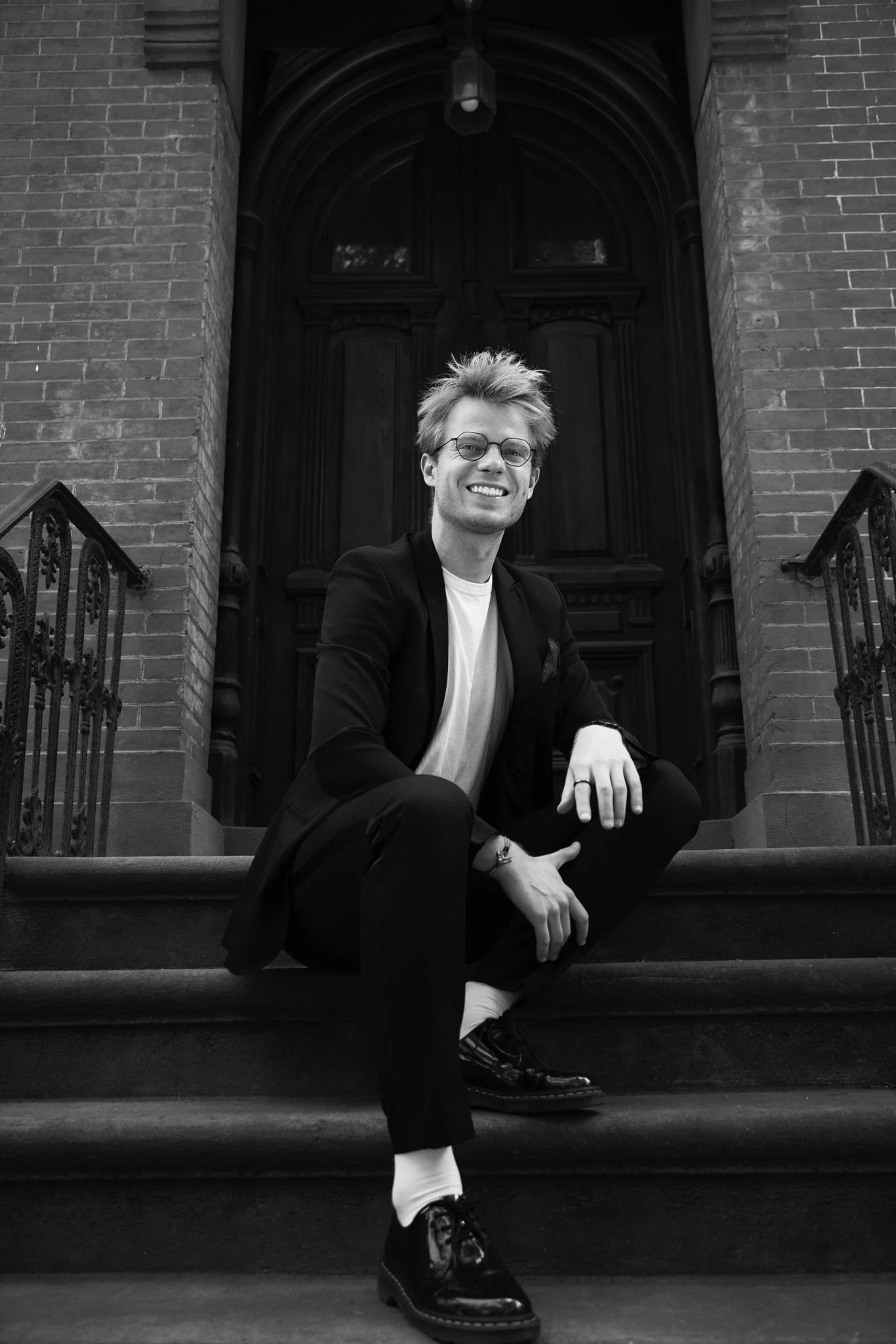 A young man with glasses, tousled hair, and a broad smile sitting on stairs outside a brick building with a large wooden door in the background.
