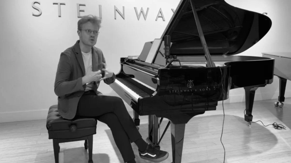 A man seated on a piano bench in front of a grand piano, in a room with the word 'STEINWAY' on the wall.