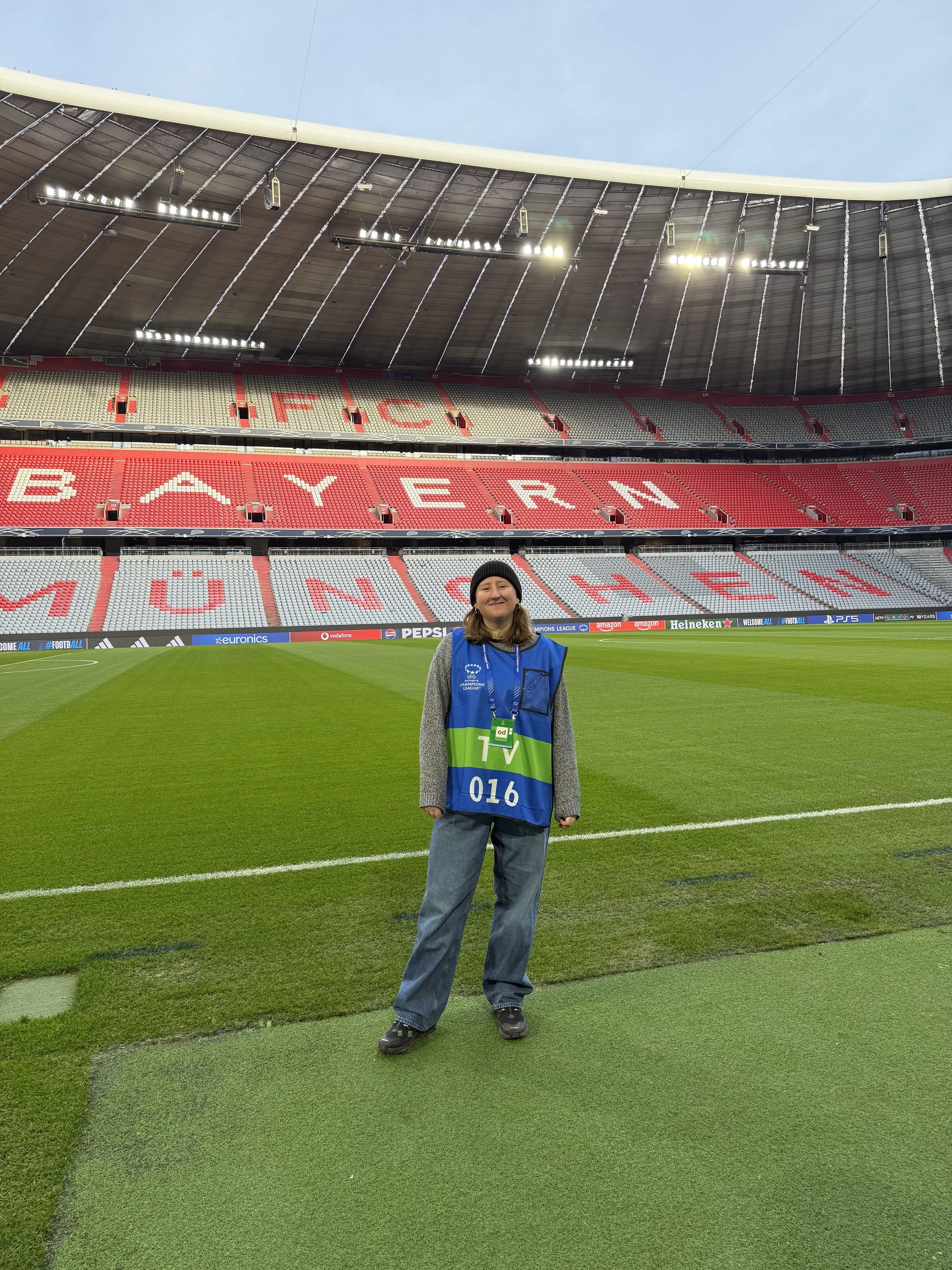 Person standing on a soccer field inside a stadium, wearing a blue vest, gray sweater, blue jeans, and a black beanie, with large empty stadium seats and a roof overhead.