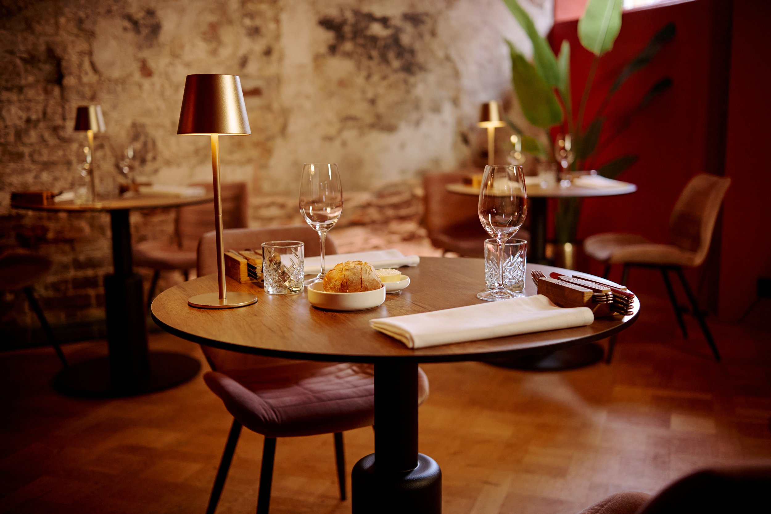 A cozy restaurant table set with a bread roll, water, wine glasses, and utensils, illuminated by a warm table lamp, with exposed brick walls and additional tables and chairs in the background.