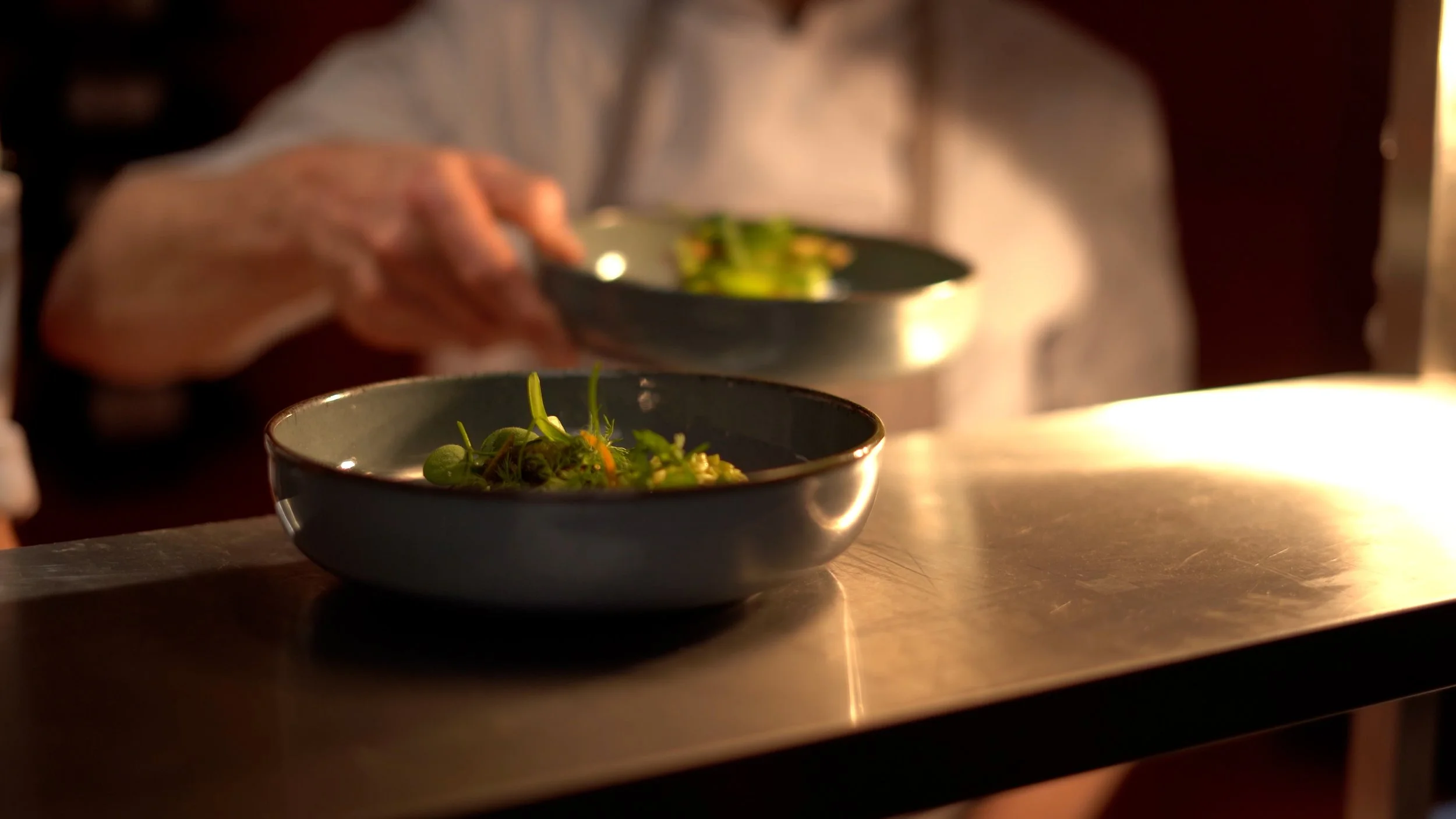 A chef or server placing small bowls of green salad or microgreens on a metal countertop in a restaurant kitchen.