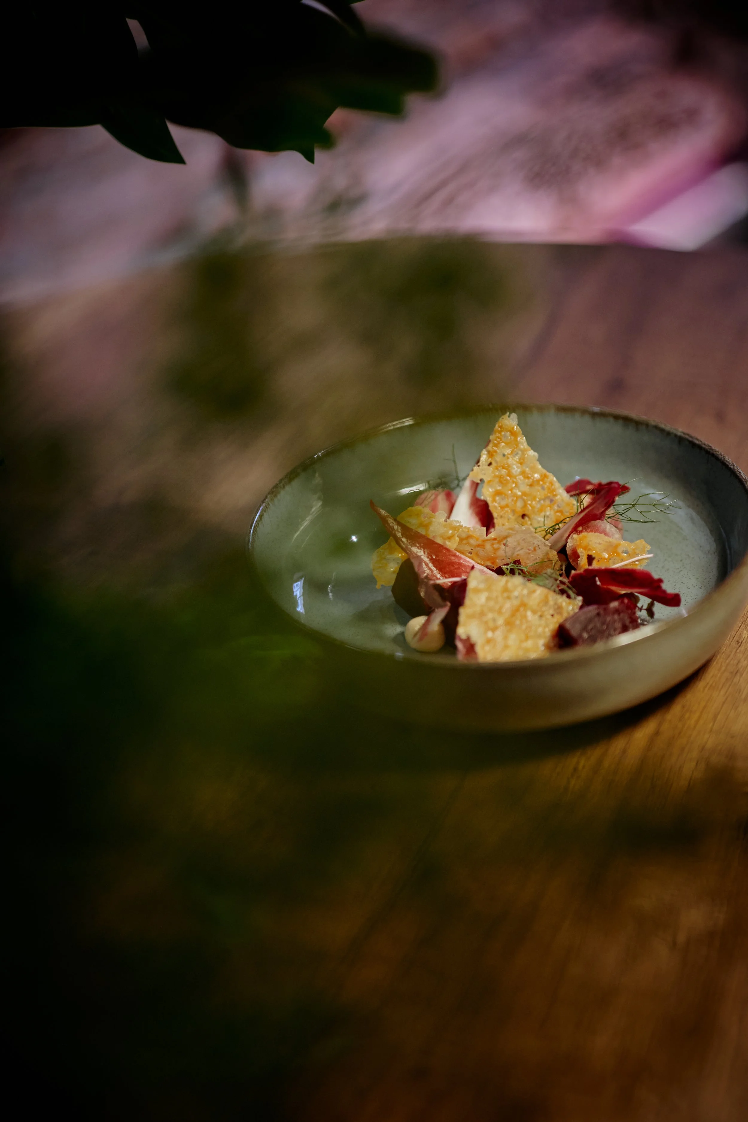 A ceramic bowl on a wooden table holds a dish with crispy chips, red leaves, and dollops of creamy sauce.