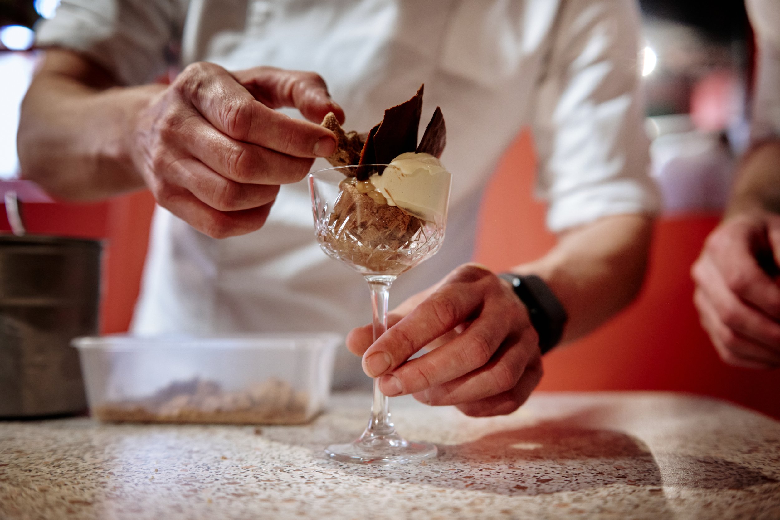 A person in a white shirt is adding chocolate to a glass dessert cup with ice cream, chocolate, and dark chocolate shavings on top. The scene is set on a speckled countertop in a restaurant or cafe.