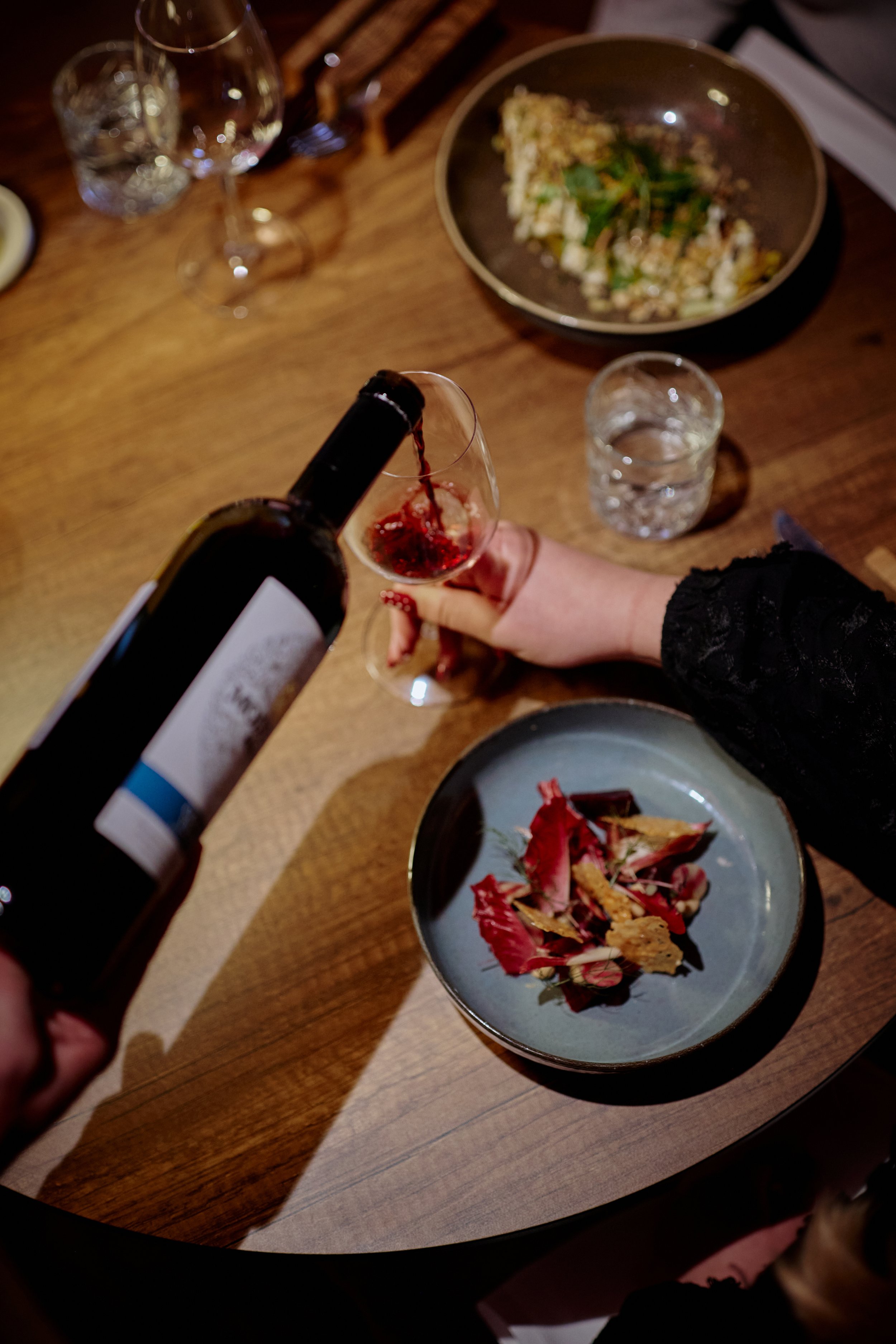 Person pouring red wine into a glass on a wooden table with plates, glasses, and food in the background.