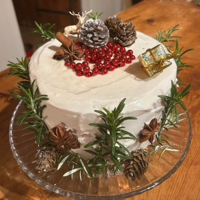 A Christmas-themed cake decorated with pinecones, red berries, star anise, cinnamon stick, a small deer figurine, and a mini wrapped gift, surrounded by green pine branches.