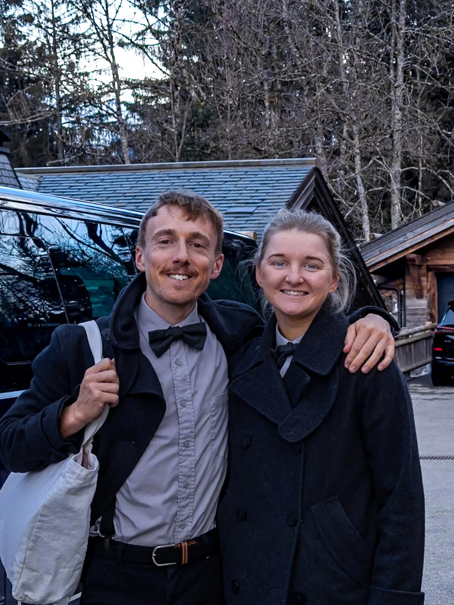 A man and woman with light skin smiling and posing outdoors, standing close together with the man's arm around the woman's shoulder. They are dressed in semi-formal clothes, with the man wearing a bow tie and the woman wearing a coat. There are trees and buildings in the background, suggesting it is late autumn or winter.