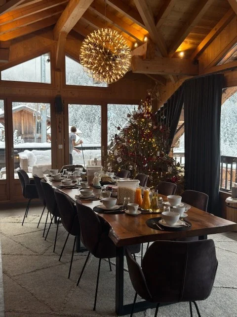 A festive Christmas dining room with a large wooden table set with white dishes, cups, and cutlery, decorated with a Christmas tree and holiday decorations, in a room with wood-paneled walls, large windows, and a mountain view outside. A chandelier h
