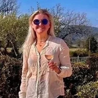 A woman standing outdoors on a brick patio, holding a glass of wine, wearing sunglasses, a light-colored jacket, black pants, and white shoes, with trees and hills in the background.