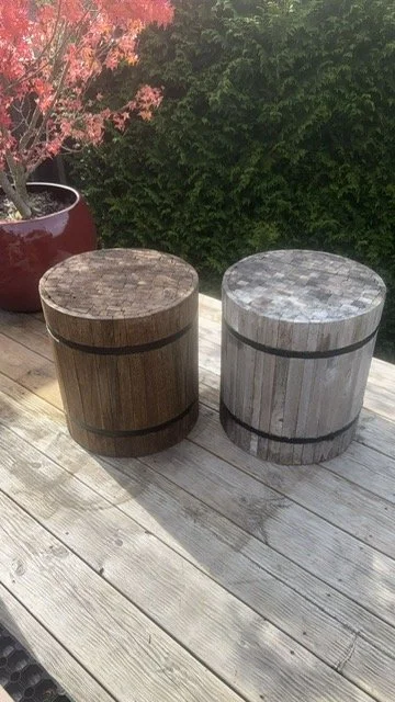 Two cylindrical wooden stools with black bands, placed on a wooden deck, with a pink flowering tree and green bushes in the background.