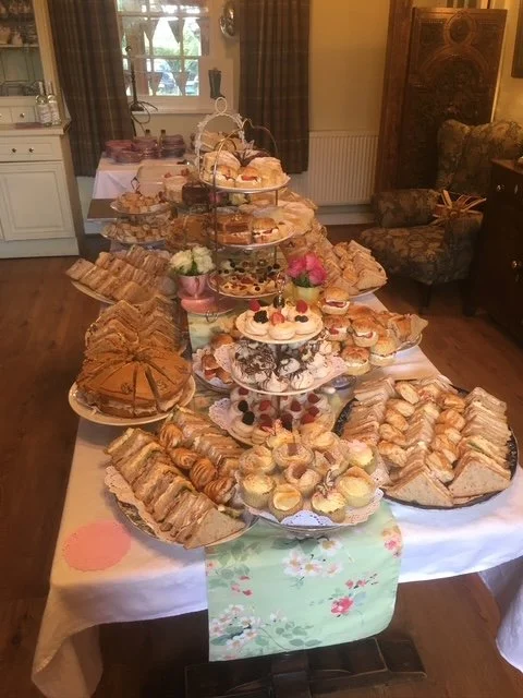 A table set with a large tiered tray of assorted cakes, pastries, and finger sandwiches in a cozy, well-lit room.