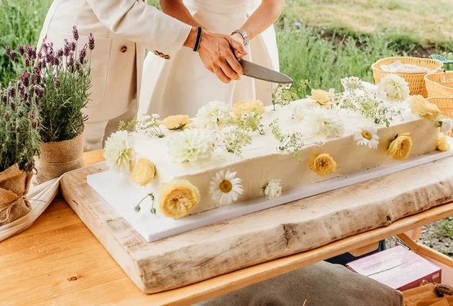 Person cutting a wedding cake decorated with white and yellow flowers, on a wooden table outdoors.