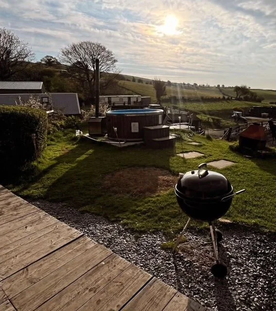 A backyard with a black kettle grill, a hot tub, a small shed, and rolling green hills in the background under a partly cloudy sky with the sun shining.