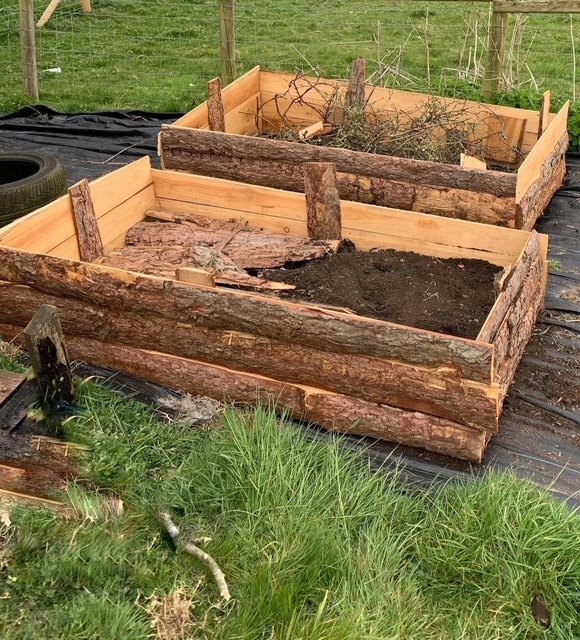 Two garden beds made of wooden logs and planks, with one filled with dark soil and the other empty. Tools and grass are visible nearby.