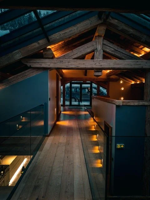 Interior view of an attic with wooden beams, skylight windows, and a hallway leading to a glass door, illuminated with warm lighting.
